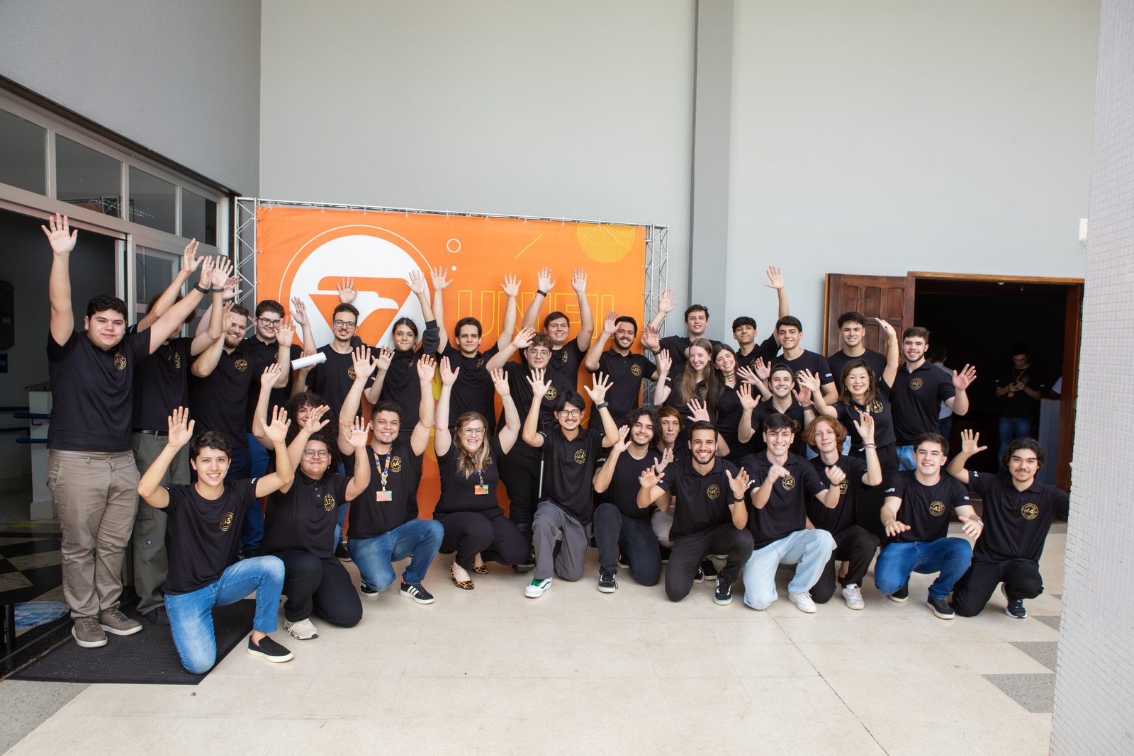 Equipe do curso de Computação da UniFil reunida, todos com as mãos levantadas, em frente a um painel laranja da UniFil.