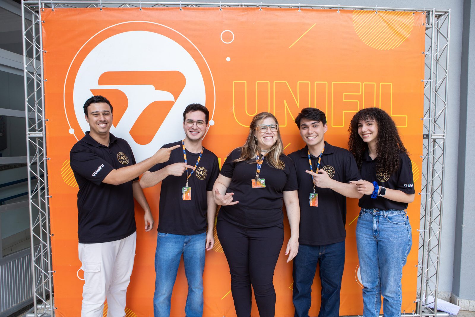Cinco integrantes da equipe da Computação UniFil posando sorrindo em frente ao painel laranja da UniFil.