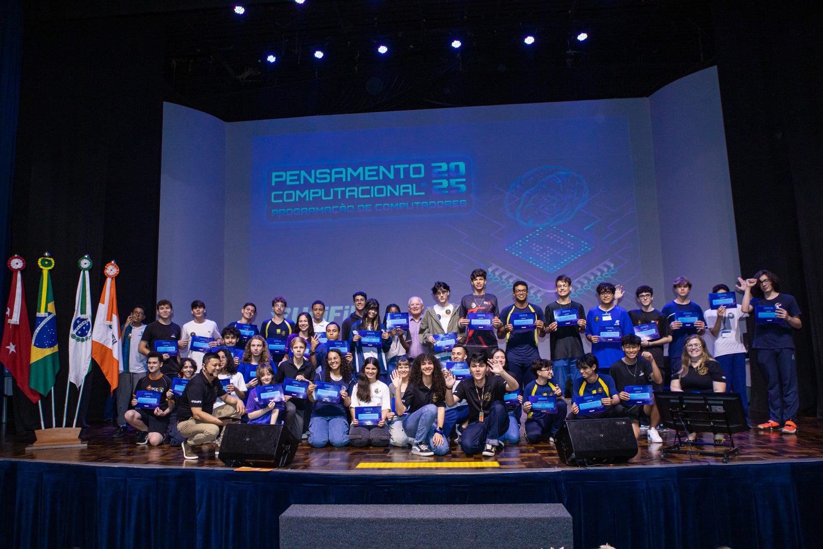 Alunos no palco segurando certificados na formatura do Pensamento Computacional 2025, com o telão do evento ao fundo.
