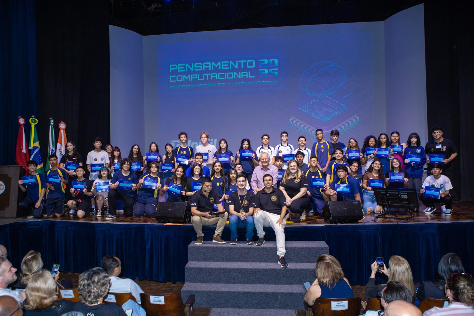 Outro ângulo da formatura do Pensamento Computacional 2025, com alunos formados e plateia em primeiro plano.