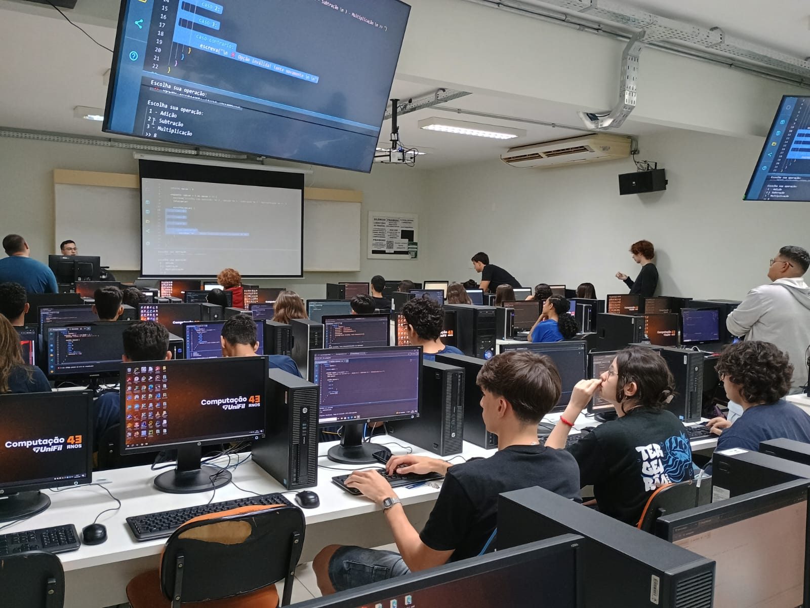 Alunos em uma aula prática de programação, sentados em computadores com exercícios de código nas telas.