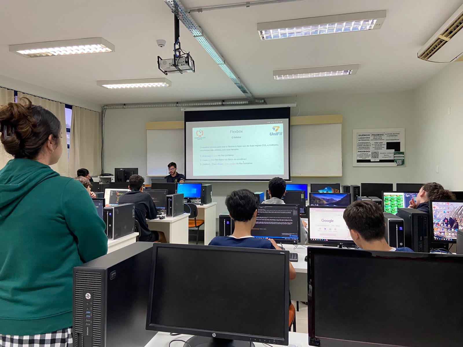 Alunos em aula prática de programação em 2026, num laboratório com vários computadores; a tela ao fundo mostra um exercício de JavaScript sobre adicionar aleatoriedade a um jogo de RPG.